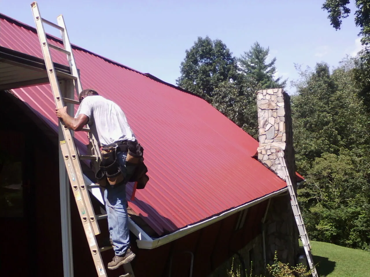 Roofer on a ladder beside a red metal roof and stone chimney in Grand Island